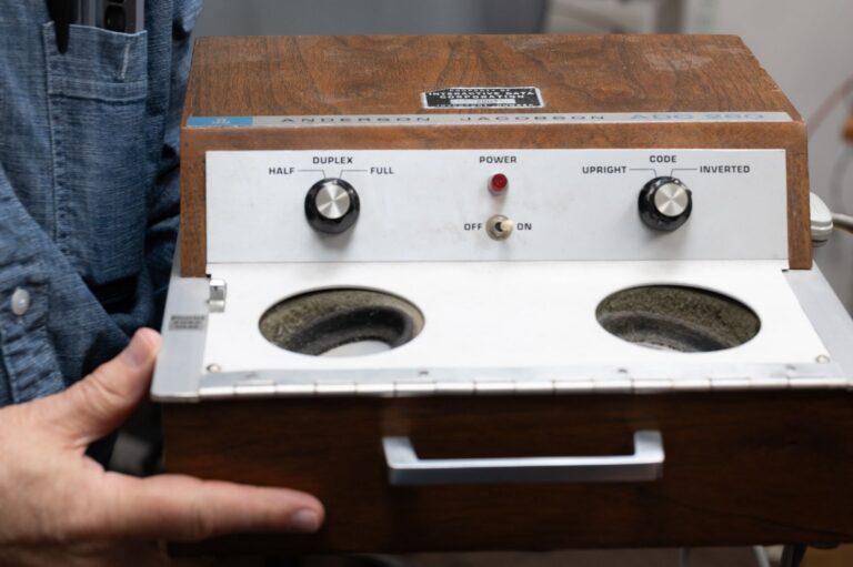 Close-up of a wood-cased Anderson Jacobson acoustic coupler with two rubber cups for a telephone handset and dials for duplex and code settings.