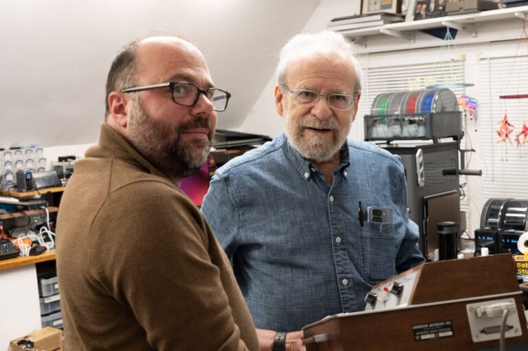 Brandon Vogel and Bob Frankston leaning over a vintage wood-grain Anderson Jacobson acoustic coupler in Bob's cluttered workshop.
