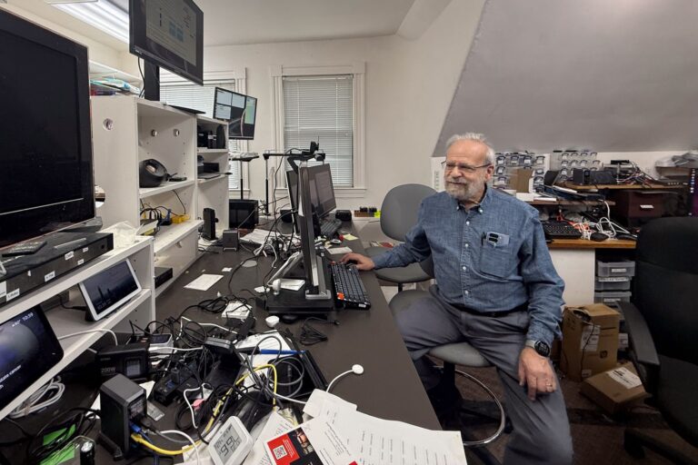 Bob Frankston seated at a long workbench in his office, surrounded by monitors, cables, and electronic gear.