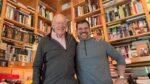 David Arscott and Aaron Sylvan standing arm in arm in front of floor-to-ceiling bookshelves in David's office.